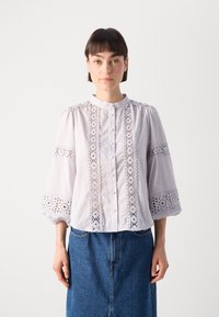 Balta blūze ar mežģīņu apdari uz piedurknēm un priekšpuses. Aprīkota ar apaļu kaklu, pogu aizdari un izpūstu piedurknēm. Valkāta ar džinsu svārkiem.