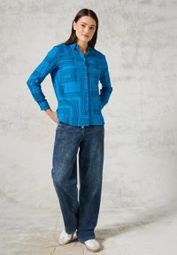 Camisa azul de botones con mangas largas que presenta un patrón geométrico de líneas negras, combinada con jeans oscuros de corte holgado y zapatillas blancas.