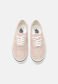 Paire de baskets Vans en daim rose clair avec lacets blancs, semelles en caoutchouc blanches et petits clous blancs près de la zone des orteils.