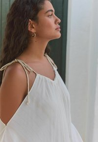 Haut blanc fluide avec des bretelles en corde, présentant une texture rayée subtile et des côtés ouverts. Des boucles d'oreilles créoles dorées ajoutent une touche métallique.