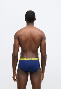 Μπλε σλιπ με κίτρινη μέση που φέρει την επιγραφή «NIKE». Ομαλό ύφασμα, εφαρμοστή γραμμή και στρογγυλεμένο σχήμα χωρίς επιπλέον σχέδια. Εμφανίζεται η πίσω όψη.