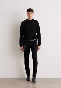 Chemise noire à manches longues et pantalon ajusté, associés à des bottines noires à la cheville. Le mannequin tient un portefeuille noir, sur un fond neutre.