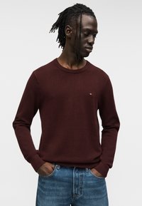 Μπορντό πουλόβερ με υφή, στρογγυλή λαιμόκοψη και μακριά μανίκια, σε συνδυασμό με ανοιχτόχρωμο τζιν παντελόνι. Μικρή λεπτομέρεια λογότυπου στο στήθος.