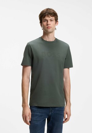 Giovane uomo che indossa una maglietta verde scuro a maniche corte con testo sottile "BOSS" e jeans blu, in piedi davanti a uno sfondo chiaro e semplice.