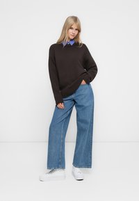 Brun strikket genser med en løs passform, blå skjorte under, parret med vide lyseblå jeans og hvite sneakers.