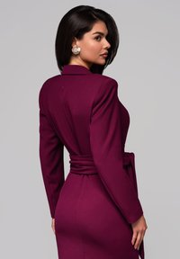 Burgundské šaty s dlouhým rukávem, splývavého siluety, s véčkovým výstřihem a páskem v pase. Hladká tkanina s elegantním zadním designem a jemnými švy.
