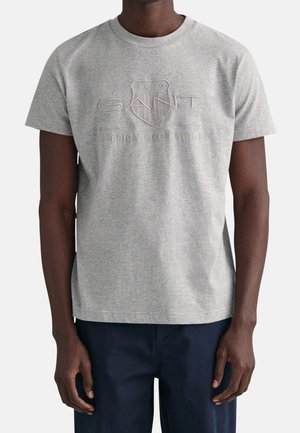 Mężczyzna ubrany w jasnoszarą koszulkę z krótkim rękawem z wytłoczonym logo "GANT 1949 American Sportswear" oraz granatowe spodnie.