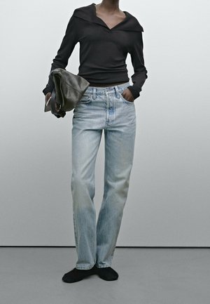 Personne portant un haut noir à manches longues avec un col large, un jean droit bleu clair, des chaussures noires, tenant une veste grise pliée.