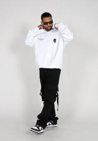 White hooded sweatshirt with front pocket, black graphic print, and black cargo pants. Model wears black sunglasses and black and white sneakers.