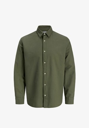 Ragazzo in piedi con le mani nelle tasche, indossa una camicia verde con bottoni, pantaloni grigi e scarpe beige su uno sfondo chiaro neutro.