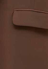 Tissu marron avec une poche à rabat visible, mettant en avant une texture lisse et un design soigné, suggérant un vêtement sophistiqué.