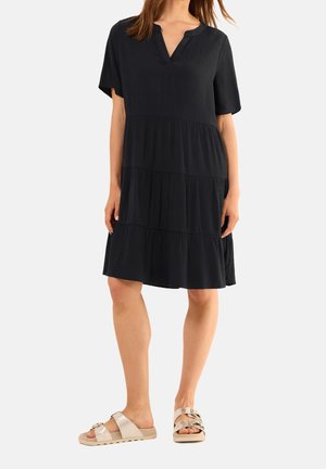 Frau trägt ein lockeres, schwarzes, knielanges, stufiges Kleid mit kurzen Ärmeln und beige flache Sandalen mit Schnallen.