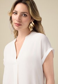 Blouse blanche avec des manches courtes retroussées, décolleté en V et en tissu léger. Possède une texture lisse et un subtil éclat. Boucles d'oreilles sculpturales en or.