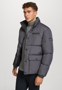 Giacca a vento grigia con colletto alto, bottoni frontali e tasche laterali. Presenta un logo sul petto e un dettaglio con zip al collo.