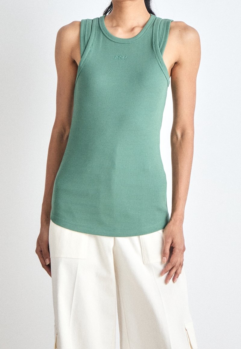 Femme portant un haut vert côtelé sans manches avec le logo "BOSS" et un pantalon blanc ample, sur fond uni.