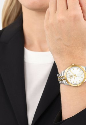Silber- und goldfarbener Armbanduhr mit weißem strukturiertem Zifferblatt, goldenen Akzenten und einem zweifarbigen Metallarmband. Tragen an einem Handgelenk in einem schwarzen Blazer.