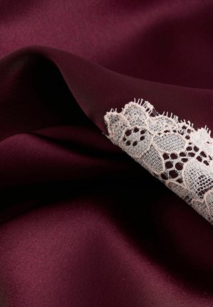Bordová saténová látka s hladkou textúrou, zdobená jemnou bielou čipkou pozdĺž okraja, ktorá zvýrazňuje zložité vzory.