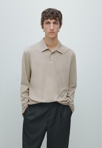 Beige poloshirt met lange mouwen, een zachte textuur en een kraag met knopen, gecombineerd met donker getailleerde broek, tegen een minimalistische achtergrond.