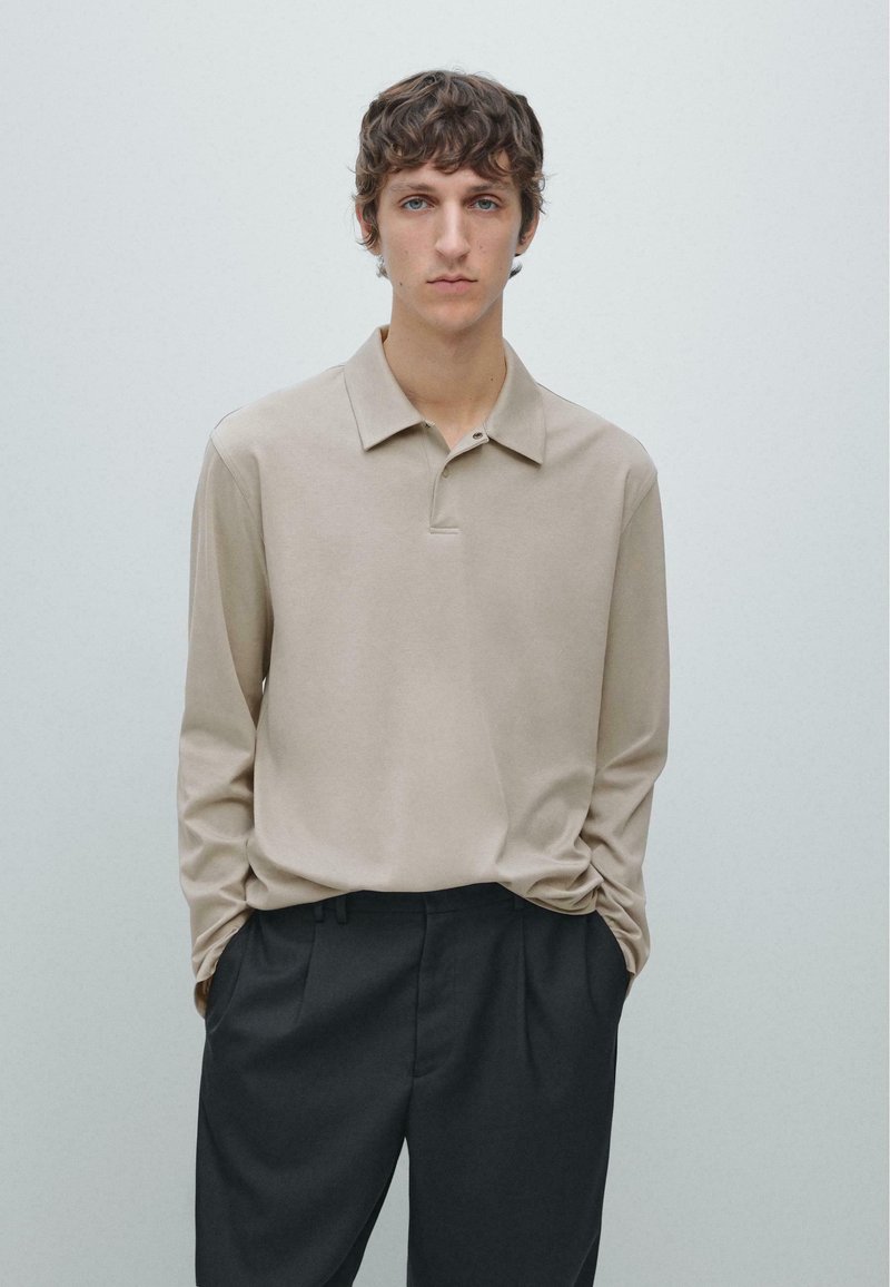 Beige poloshirt met lange mouwen, een zachte textuur en een kraag met knopen, gecombineerd met donker getailleerde broek, tegen een minimalistische achtergrond.