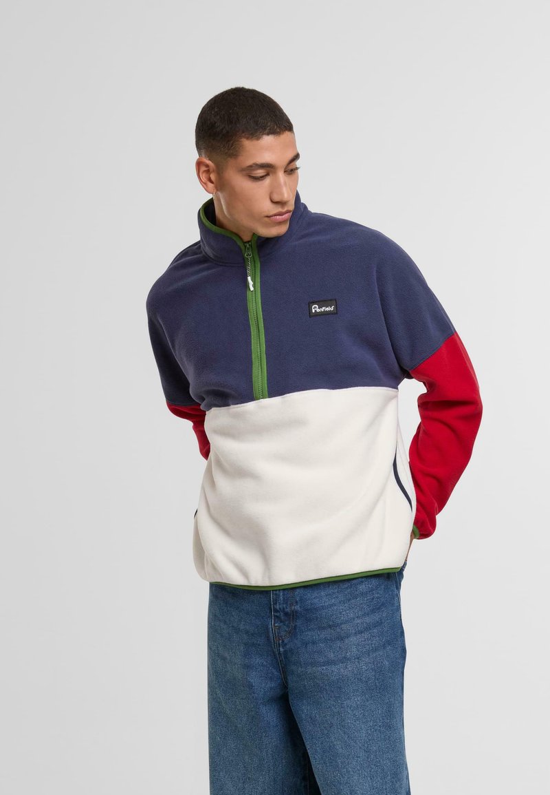 Pulover din fleece cu un corp albastru închis, mânici roșii și o secțiune inferioară crem. Fermuar verde și patch cu logo pe piept. Textură moale.