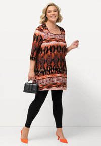 Tunic multicolore con motivo geometrico, maniche corte, abbinata a leggings neri e décolleté arancioni. Tiene una borsa nera.