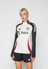 Fehér, hosszú ujjas mez piros és tengerészkék díszítéssel, negyed cipzáras gallérral, Manchester United logóval és "Tezos" felirattal. Fekete rövidnadrág is tartozik hozzá.