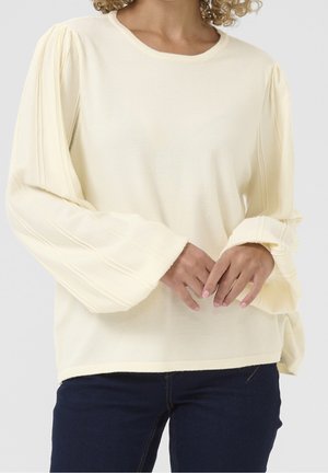 Kvinde iført cremefarvet sweater med brede, plisserede lange ærmer og mørkeblå jeans, hænderne foldet foran.