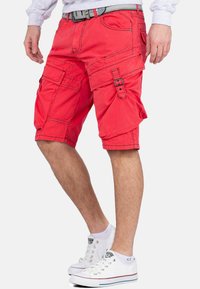 Rode cargoshorts met meerdere zakken, verstelbare banden en stikdetails. Draagt met witte sneakers met gekleurde accenten.