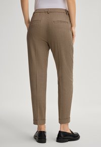 Pantalons à carreaux bruns avec une coupe ajustée, ourlets retroussés et deux poches arrière, associés à des mocassins noirs, sur un fond clair.