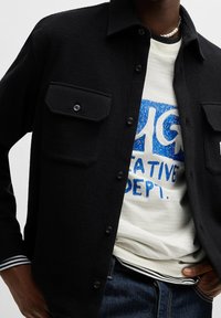 Schwarze, strukturierte Hemdjacke mit zwei Brusttaschen, Knopfverschluss und einem kontrastierenden gestreiften Bündchen, über einem cremefarbenen T-Shirt mit Grafik.