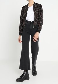 Femme portant une veste bomber noire à fleurs, un t-shirt blanc, un jean noir évasé taille haute, et des bottines noires à lacets.