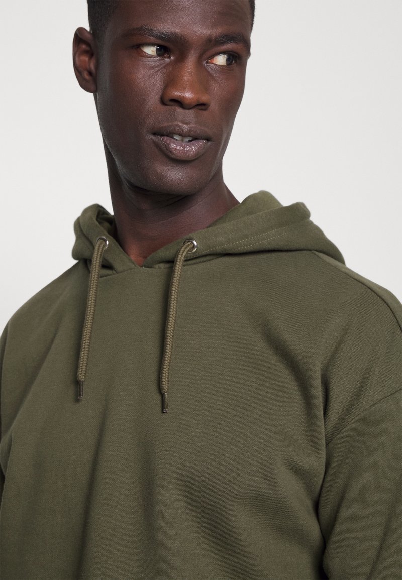 Oliwkowa bluza z kapturem z regulowanym ściągaczem, wykonana z miękkiego materiału, z metalowymi akcentami na ściągaczach i luźnym krojem.