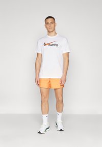 Camiseta blanca de manga corta con un logo negro y naranja de "Nike Running", combinada con pantalones cortos naranjas, calcetines blancos y zapatillas deportivas.