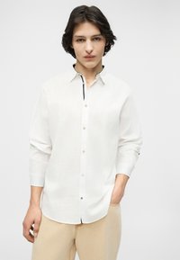 Νεαρός άνδρας με σκούρα μαλλιά που φοράει λευκό πουκάμισο με κουμπιά και μπεζ παντελόνι, στέκεται μπροστά σε ένα απλό φωτεινό φόντο.