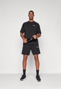 Conjunto atlético preto composto por uma camisola de mangas curtas e calções. Apresenta um logótipo na camisola, e uma pessoa a segurar uma garrafinha de água preta.