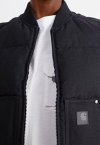 Černá bomber bunda se zapínáním na zip, žebrovaným límcem a dvěma předními kapsami. Obsahuje šedou nášivku s logem na levé kapse. Nohou ji přes bílou košili.