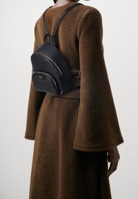 Persoon in een bruine fuzzy jas met brede mouwen en een kleine zwarte leren rugzak met gouden ritsen op hun rug.