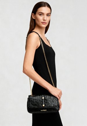 Donna con vestito nero senza maniche che porta una borsa a tracolla in pelle nera con motivo a rilievo, con catenella dorata e dettaglio logo.