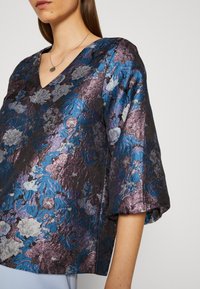 Haut floral violet avec un décolleté en V, en tissu métallique, avec des motifs de fleurs texturés et des manches courtes et bouffantes.