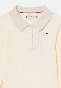 Krémový pletený polo svetričok so strieborným golierom. Obsahuje káblovú textúru, tri gombíky pri krku a malé logo na hrudi.