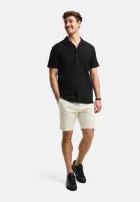 Black short-sleeve button-up shirt, cream shorts, black sneakers. Smooth fabric, relaxed fit, and buttons down the front. Simple design.