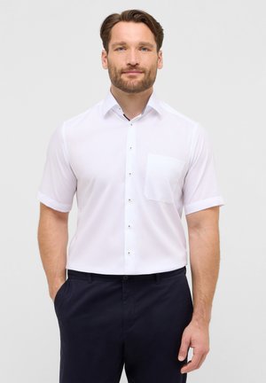 Weißes Kurzarmhemd mit Button-down-Kragen, ausgestattet mit einer einzelnen Brusttasche und kontrastierenden Knöpfen, kombiniert mit dunkelblauen Hosen.