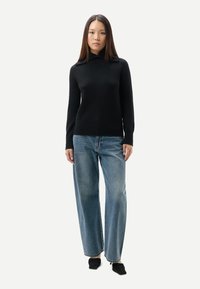 Svart stickad tröja med hög krage, kombinerad med ljusblå vida jeans. Modellen bär svarta slip-on skor. Enkel och ren design.