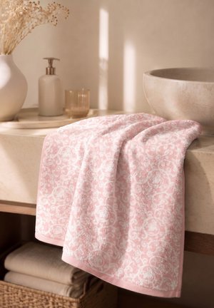Serviettes roses douces à motifs, drapées sur un comptoir de lavabo en pierre beige avec un distributeur de savon, un vase de fleurs séchées et des serviettes pliées en dessous.