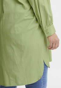Camicia a maniche lunghe verde chiaro con polsini abbottonati, presenta un orlo curvo e una texture morbida. Abbinata a jeans blu.
