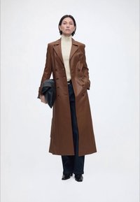 Γυναίκα όρθια φορώντας μακρύ καφέ δερμάτινο παλτό, κρεμ ζιβάγκο, σκούρο παντελόνι, μαύρα παπούτσια και κρατώντας μια μαύρη τσάντα χειρός.