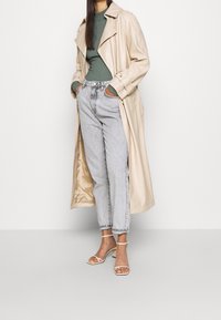 Vrouw in een beige lange jas, groene ribbeltrui, lichtgrijze jeans en witte gekruiste haksandalen, staande met haar handen in de zakken.