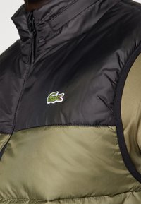Gilet rembourré noir et vert olive avec un col haut, arborant le logo crocodile de Lacoste. Texture lisse avec des matériaux contrastants.