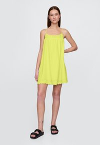 Neon rumena obleka brez rokavov z čipkastim robom na robu, lahka tkanina, dolžine do kolen, nošena s črnimi sandali.