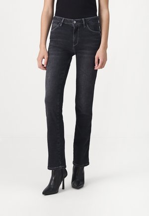 Persoon die zwarte recht-fit jeans en zwarte enkellaarsjes met een puntige neus en hoge hak draagt, staand tegen een effen witte achtergrond.